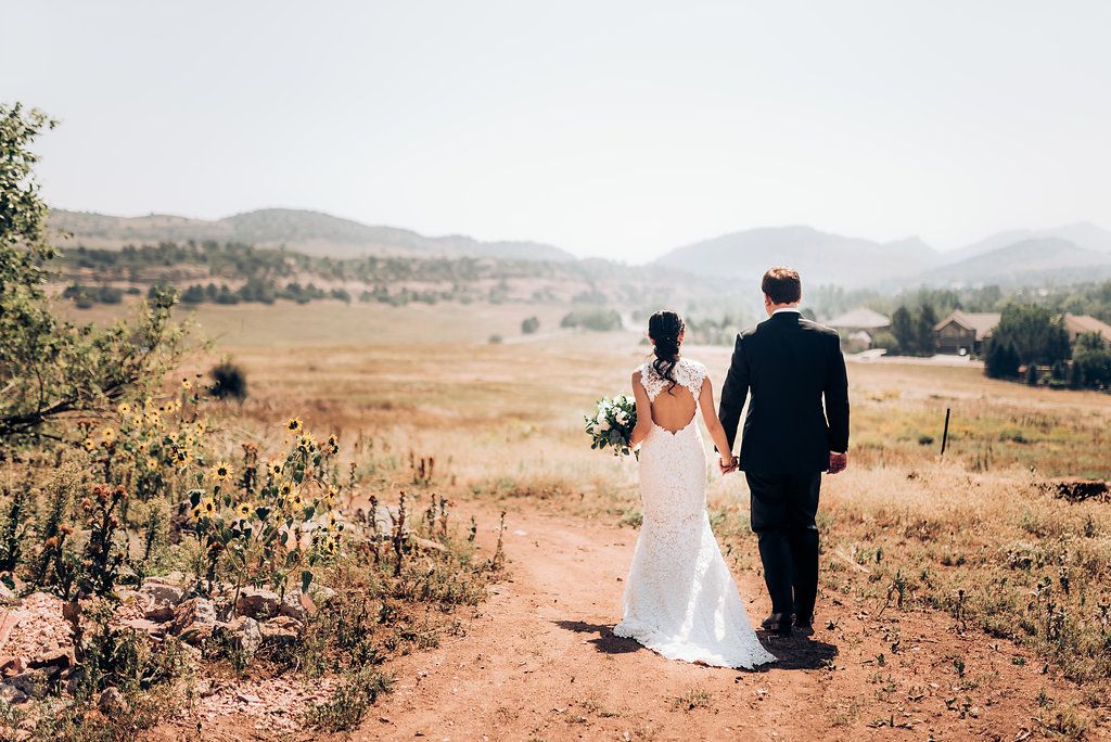 Couple walks hand in hand through golden field with mountains in the distance.