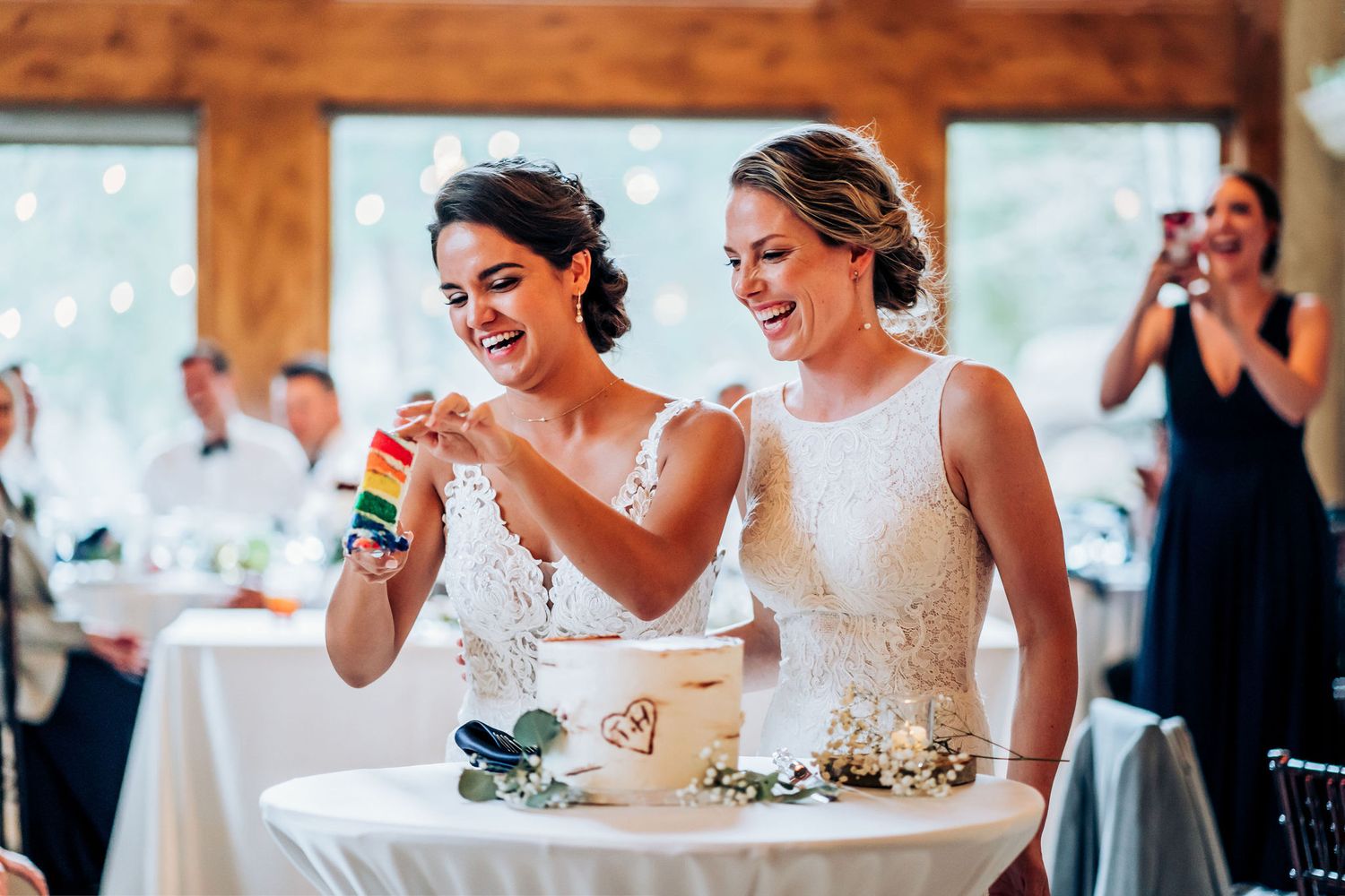 Two people in lace white dresses smile joyfully while cutting their wedding cake at an indoor reception.