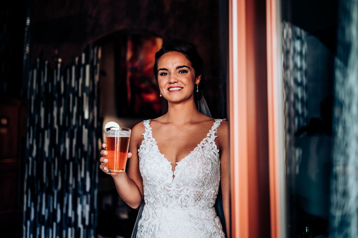 A radiant bride in a lace V-neck wedding dress holds a glass of beer.