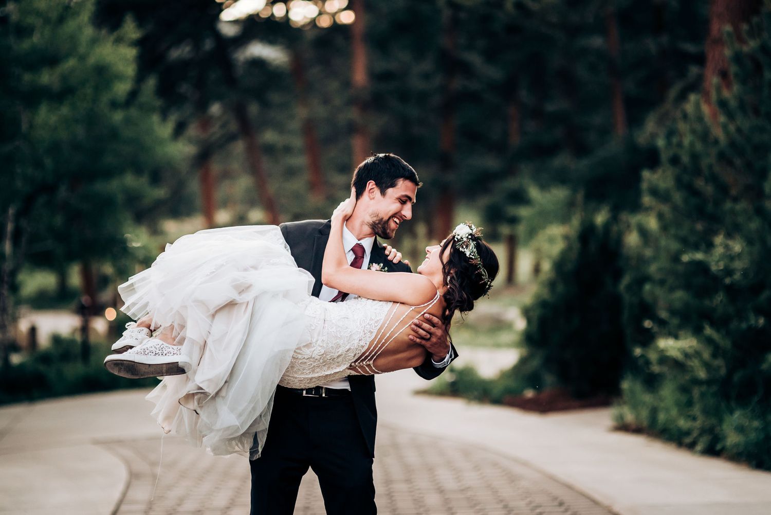 Groom dips bride in romantic pose along tree-lined pathway in classic wedding attire.