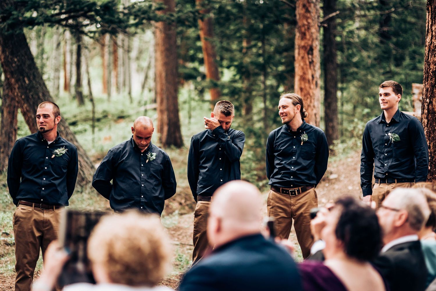 Groomsmen in matching navy shirts stand in formation during a forest wedding ceremony.