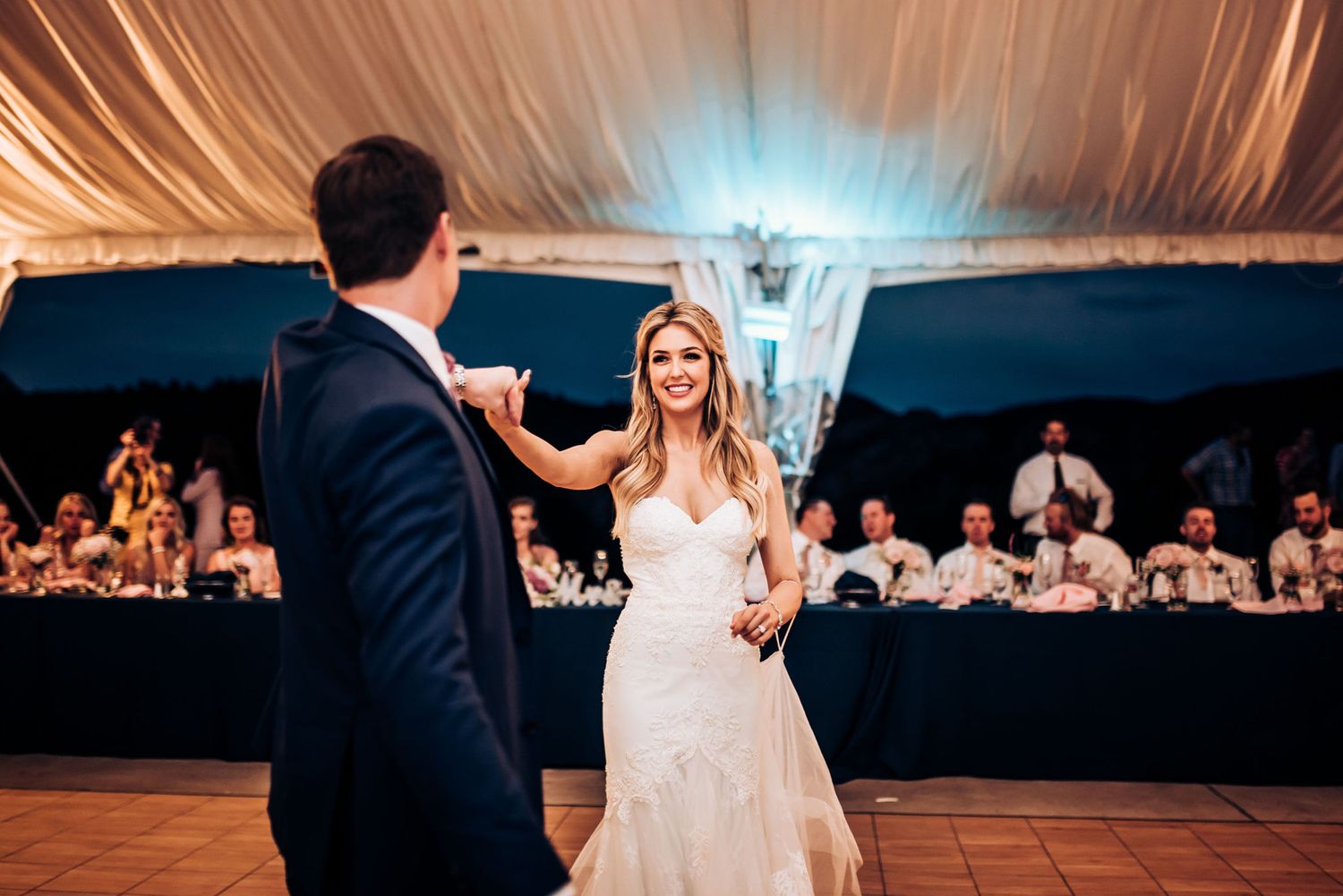A couple shares their first dance at a wedding reception in a beautifully draped tent venue with turquoise lighting.