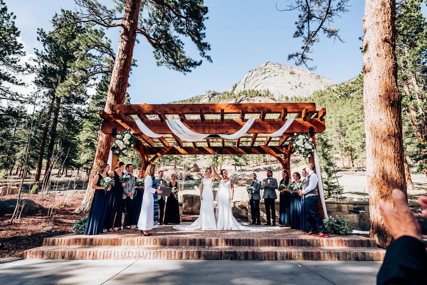 A wide view of a wedding ceremony under a wooden pergola with pine trees and mountains in the background.