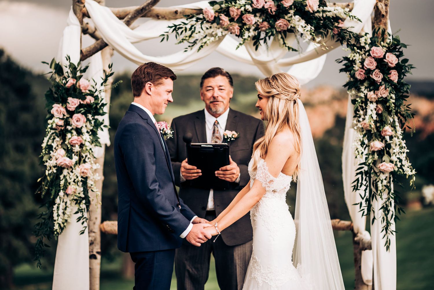 Wedding ceremony under a white arch decorated with pink and white flowers and greenery.