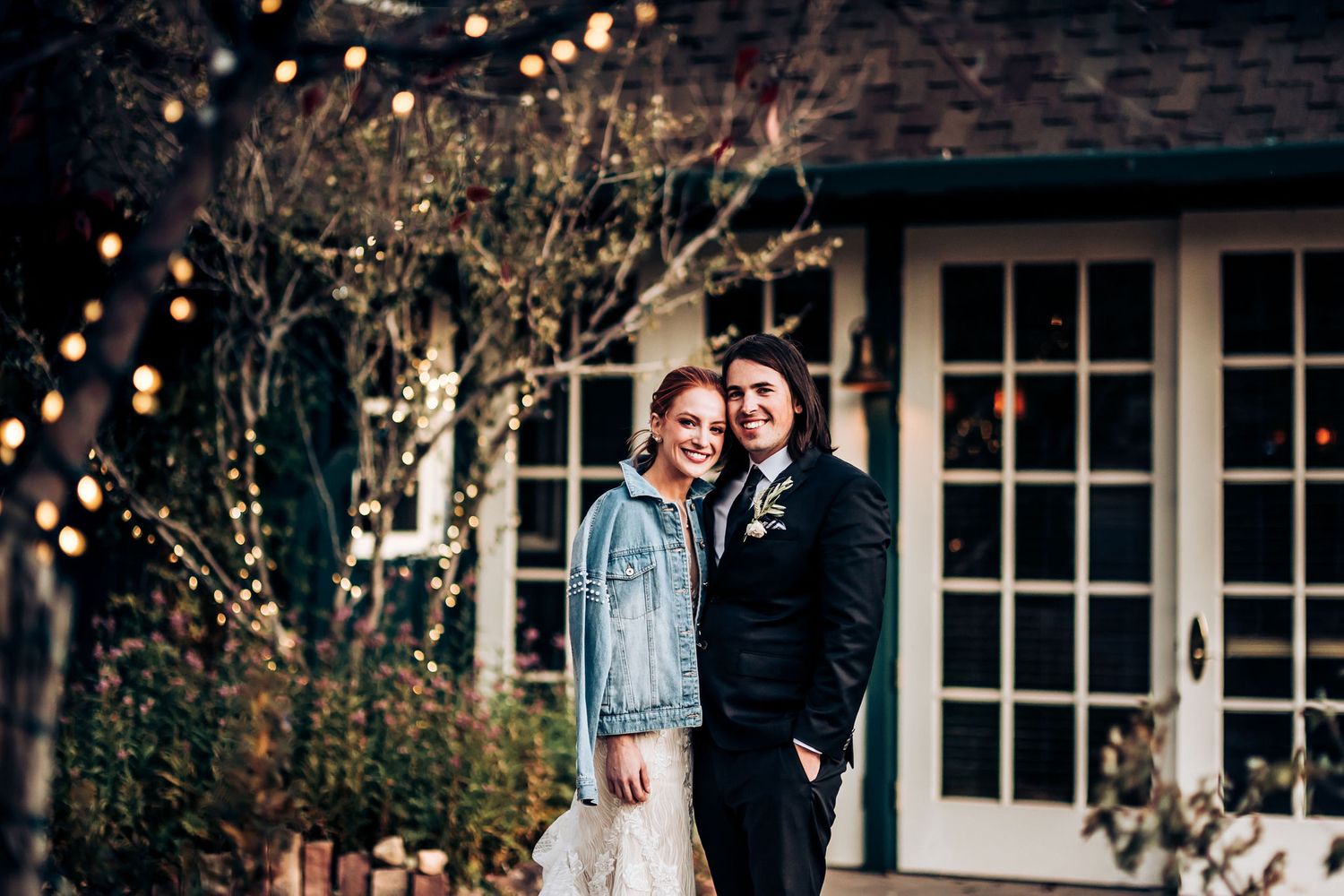 A romantic wedding couple poses in front of a brick building with twinkling string lights at dusk.