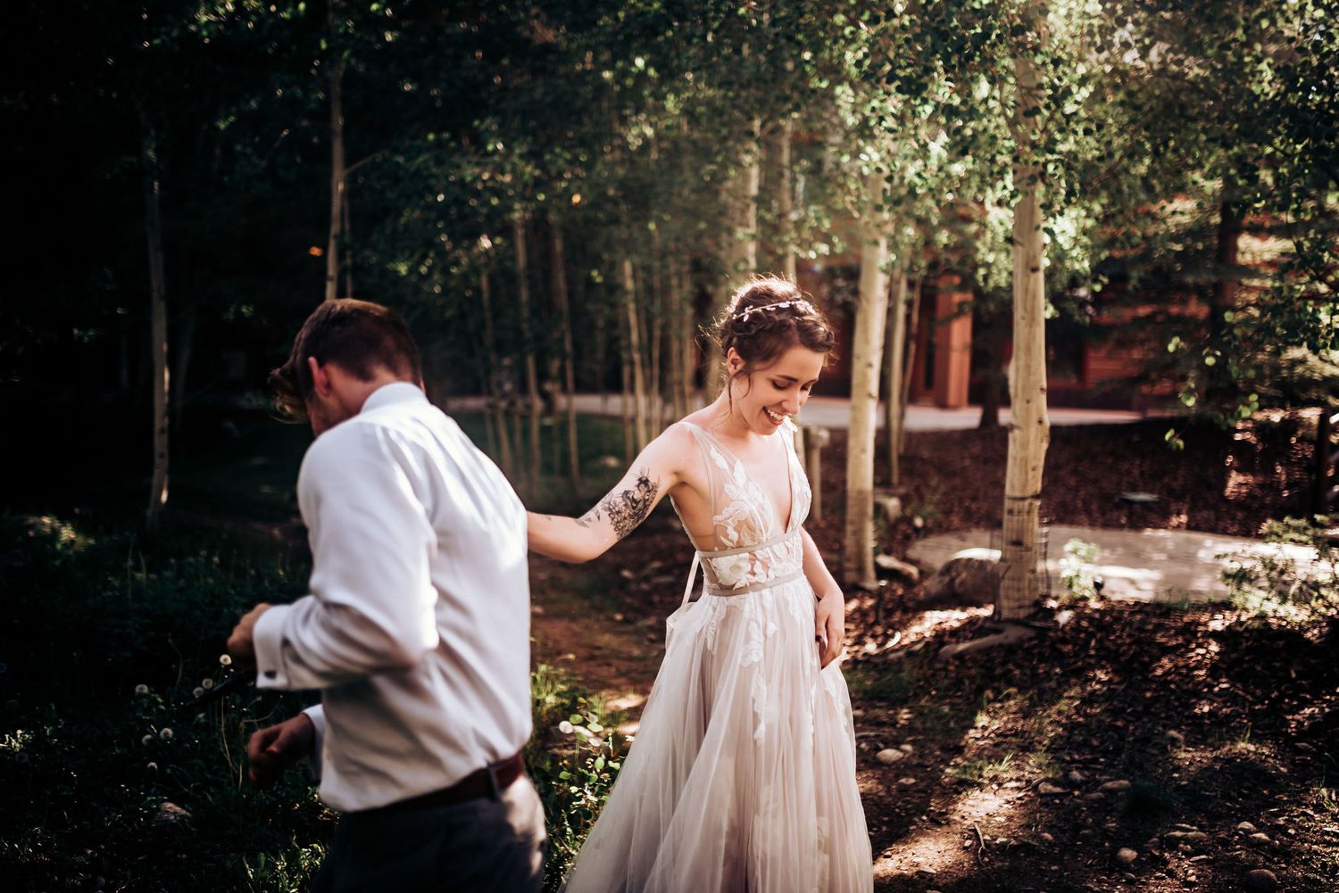 A couple in wedding attire poses in a wooded setting with dappled sunlight filtering through trees.