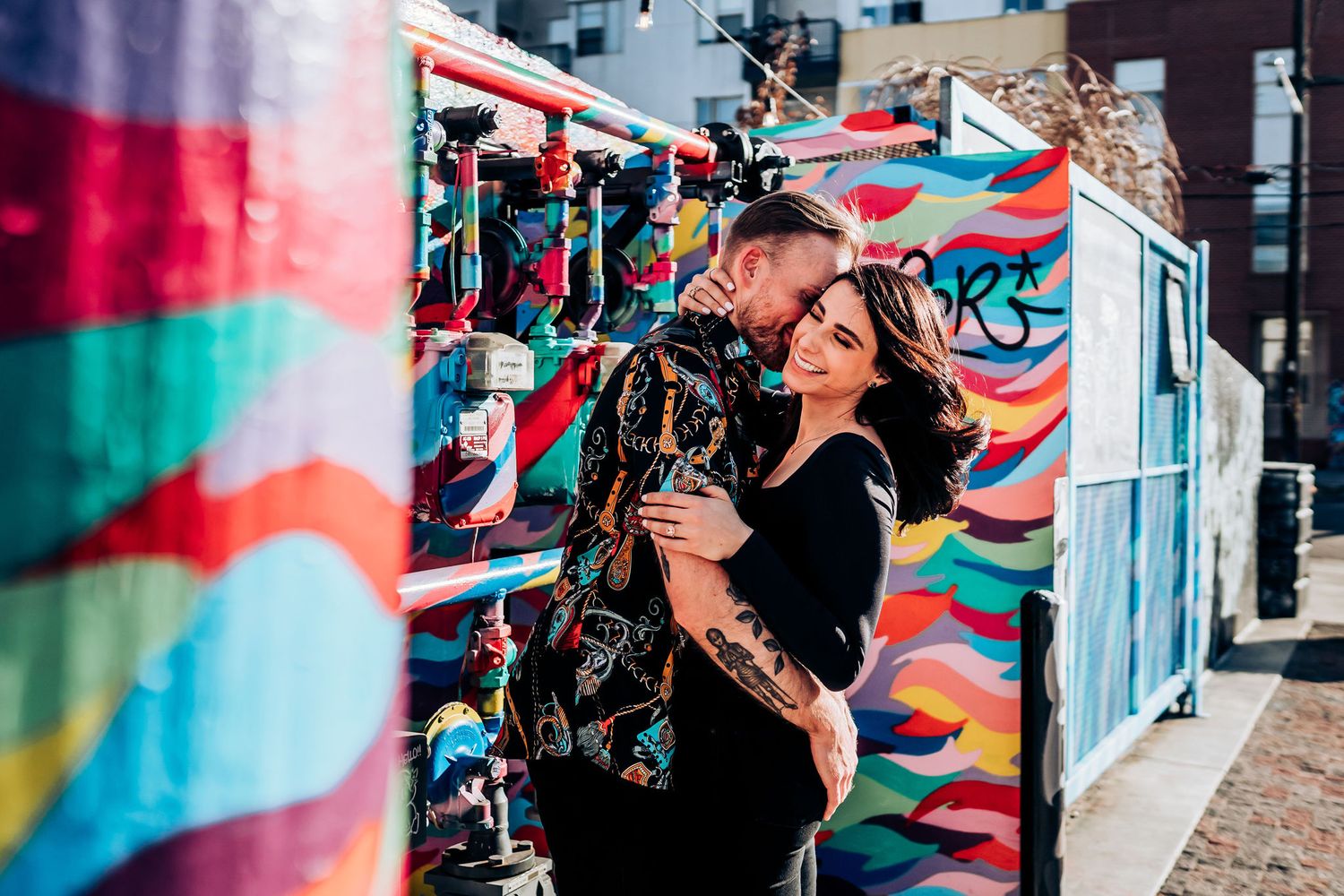 Couple embraces against colorful street art mural wall with bright geometric patterns and designs.