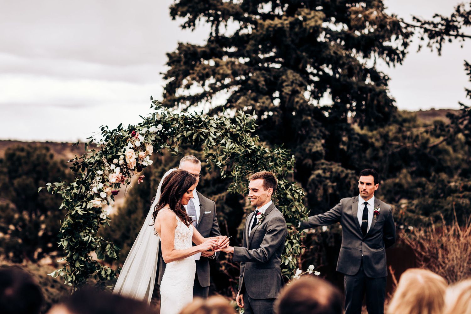 Couple exchanges wedding vows under a floral arch during an outdoor mountain ceremony with rustic backdrop.
