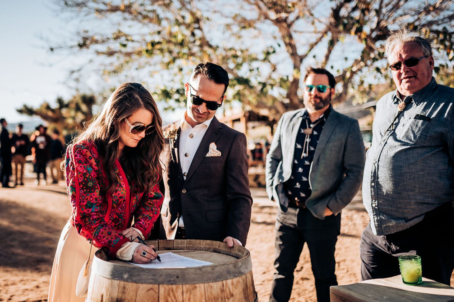 Wedding guests gather around a rustic wooden barrel for document signing in an outdoor desert setting.