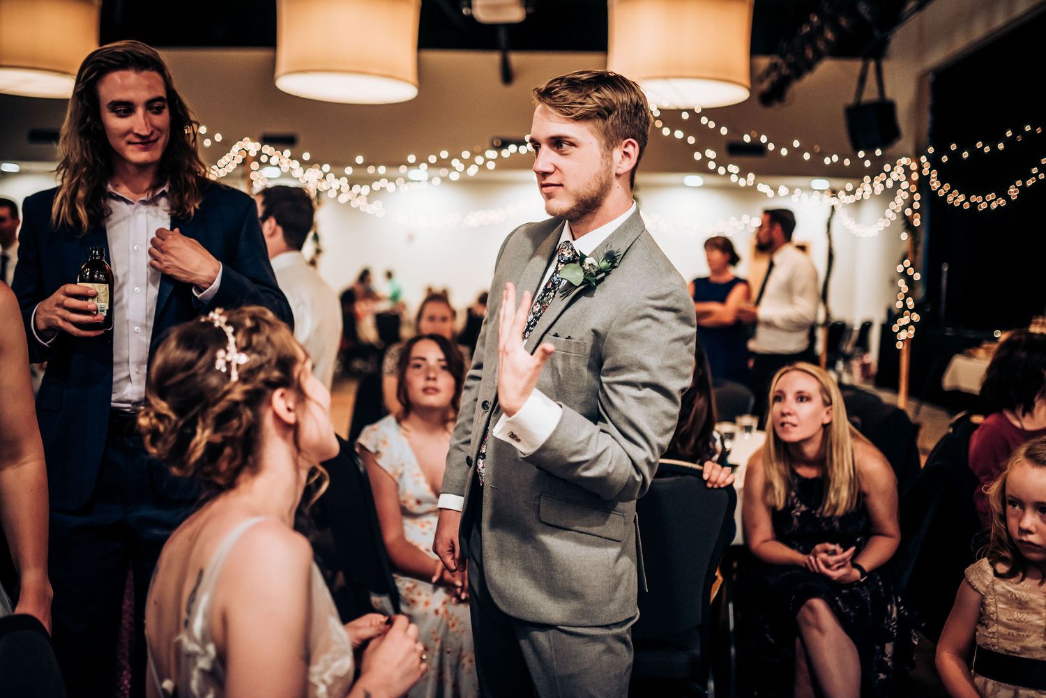 Wedding guests gather at a reception venue decorated with string lights as a person in a grey suit speaks.
