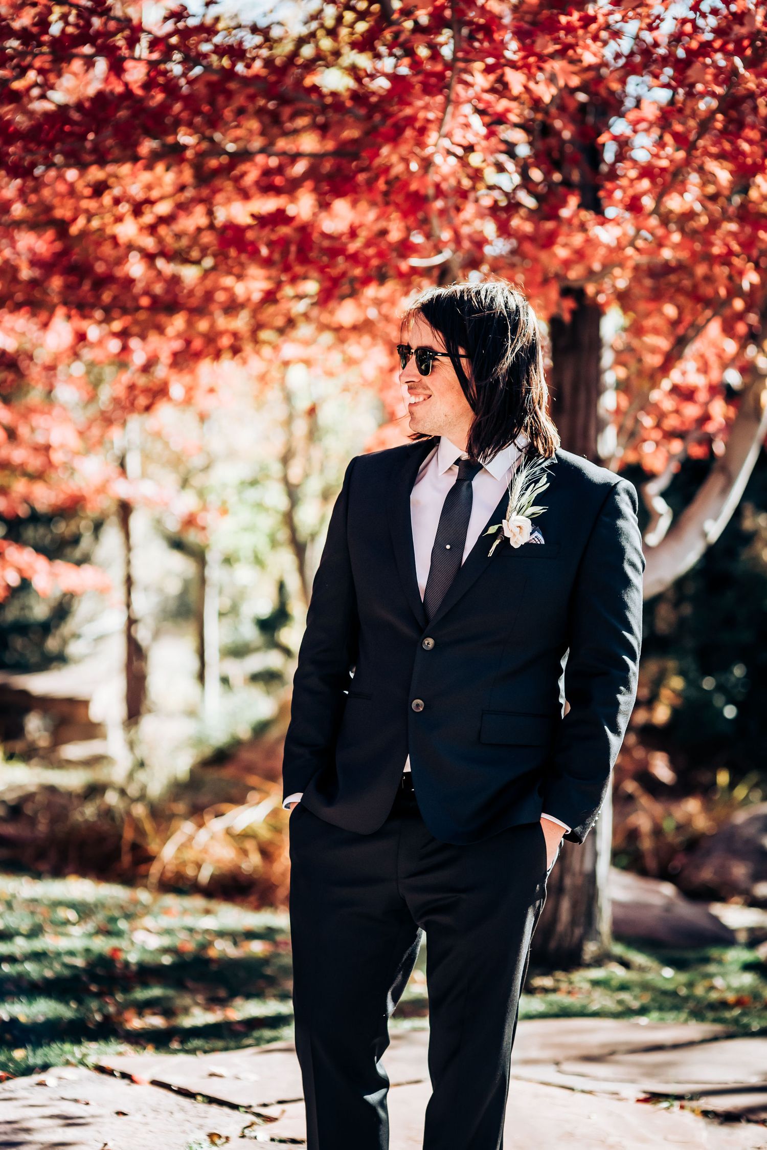 Stylish wedding attire with black suit and sunglasses against autumn foliage backdrop.