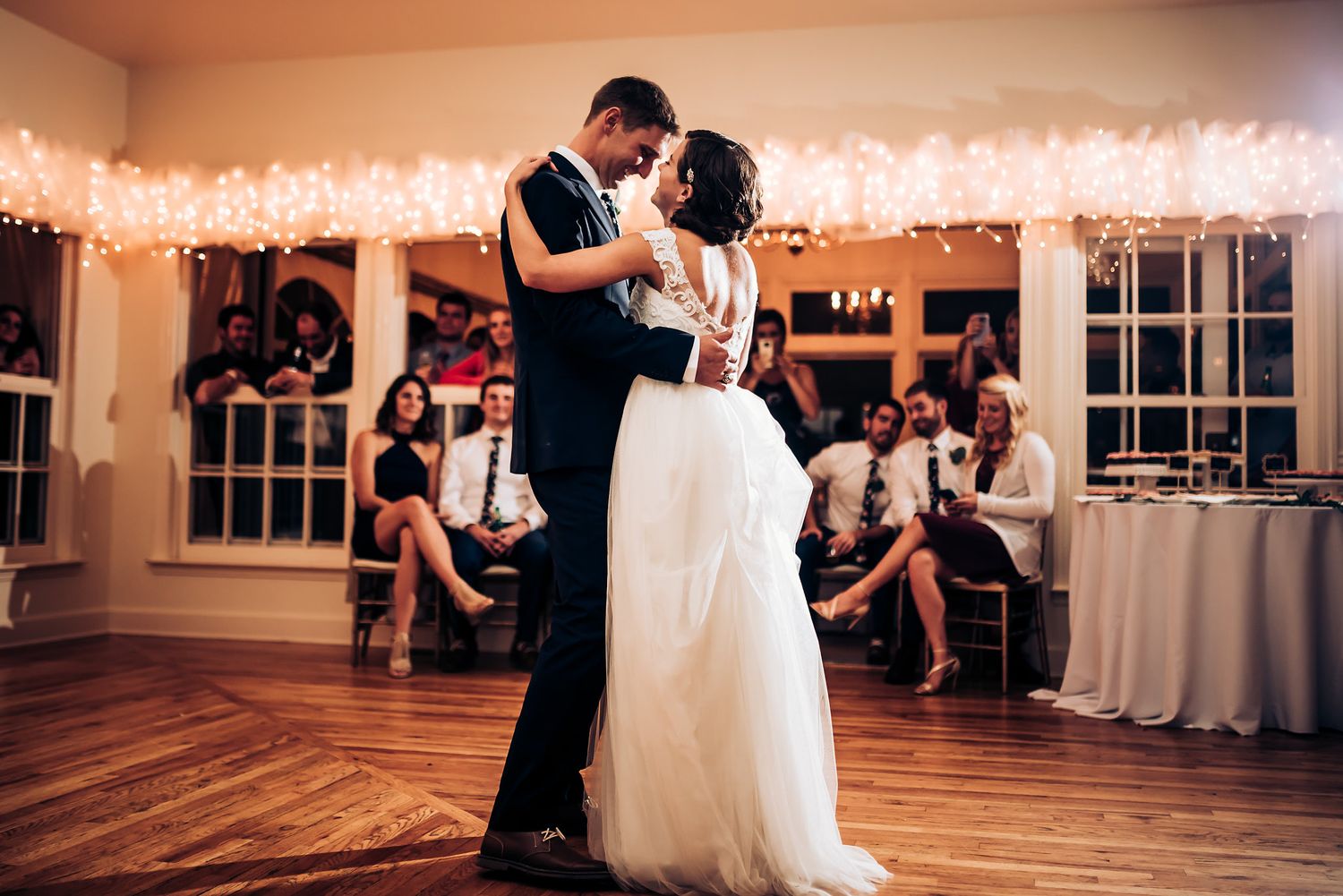 Newlyweds share their first dance under string lights while guests watch from the sidelines.