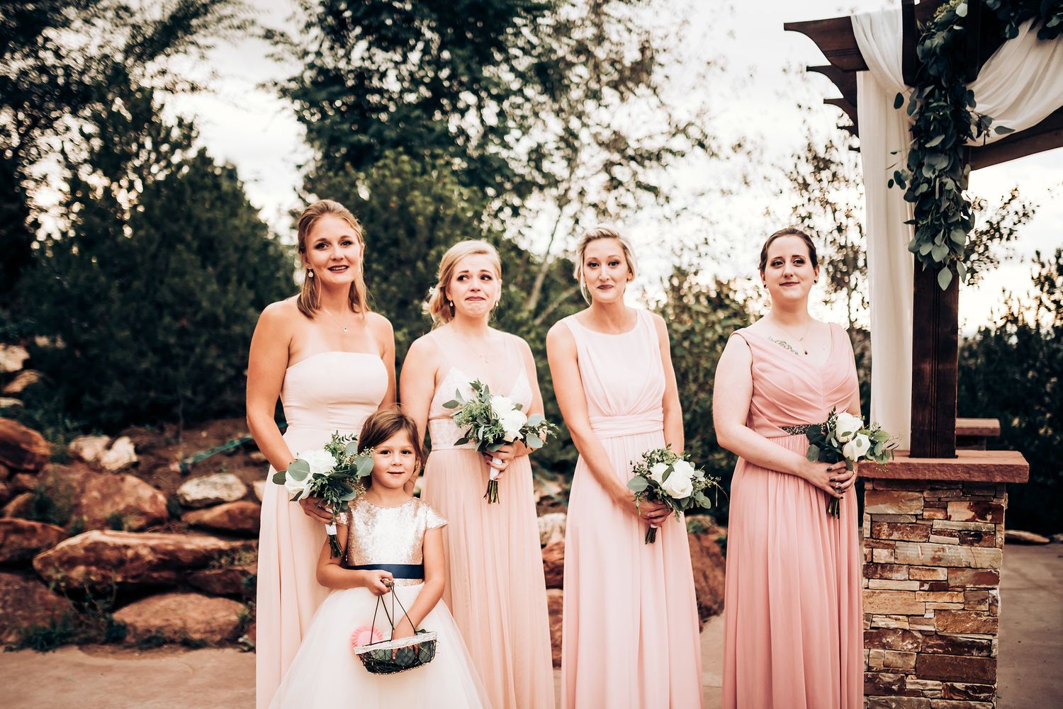 Bridesmaids in blush pink gowns stand together holding bouquets under a decorated wedding arch.
