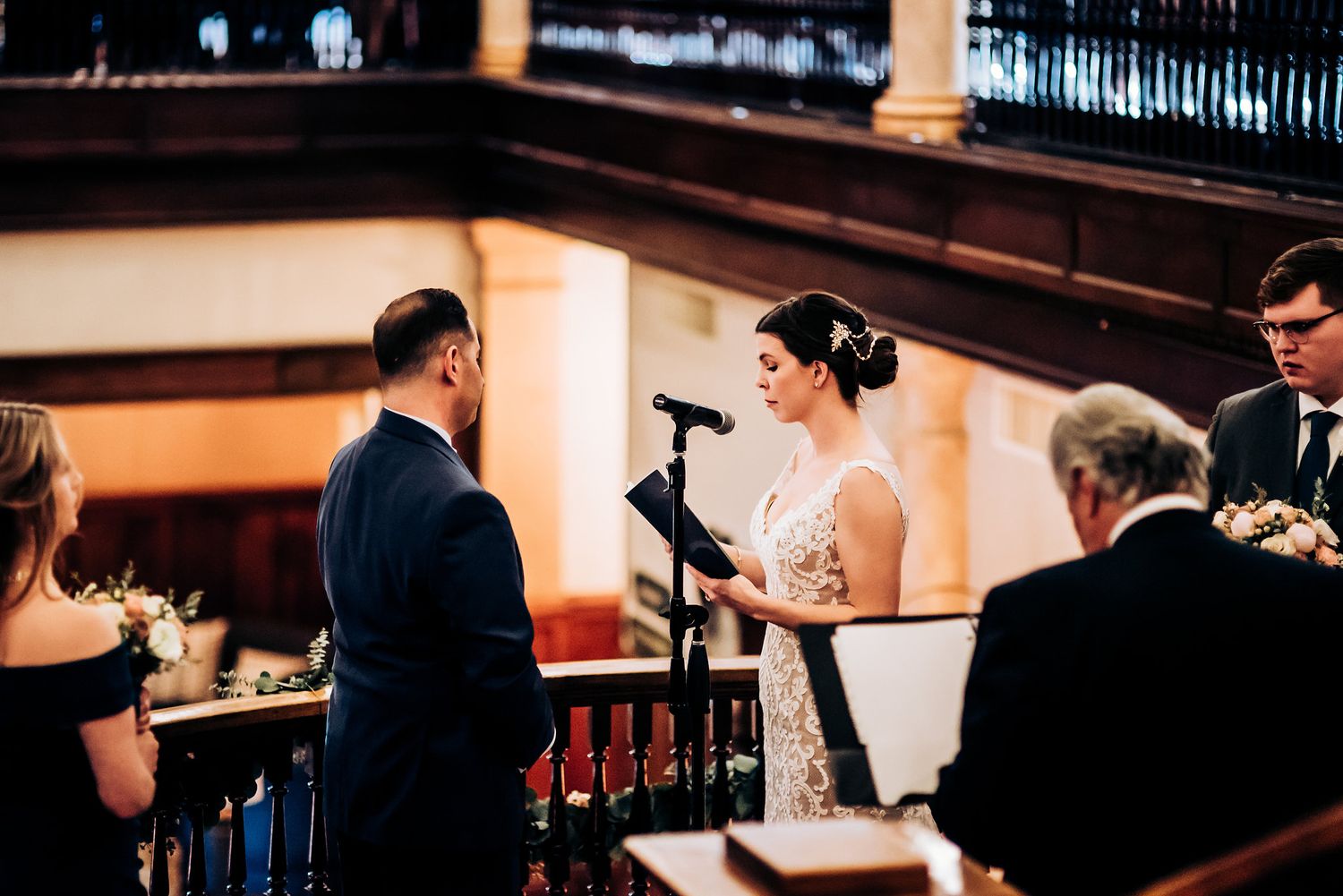 An intimate wedding ceremony progresses with participants exchanging vows in a historic interior setting.