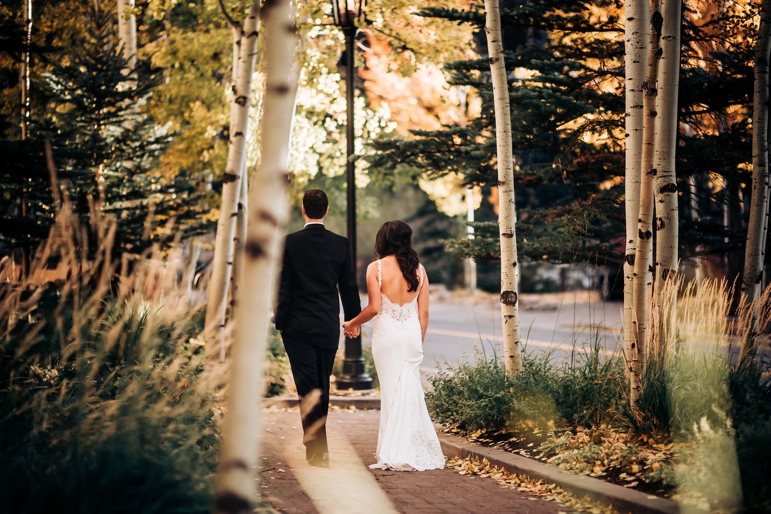 A couple walks along a scenic path through birch trees at sunset during their wedding photoshoot.