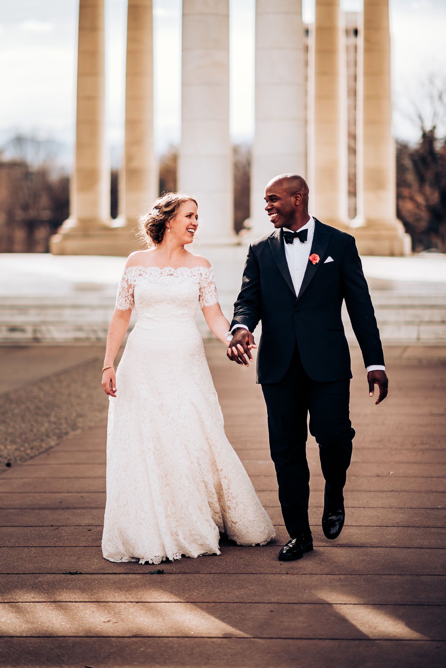 Newlyweds walk together holding hands among classical white pillars.
