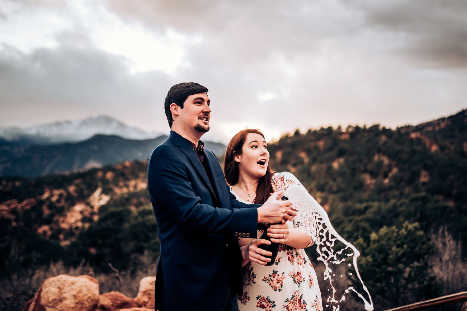 A couple laughs together while standing on red rocks overlooking a mountain landscape.