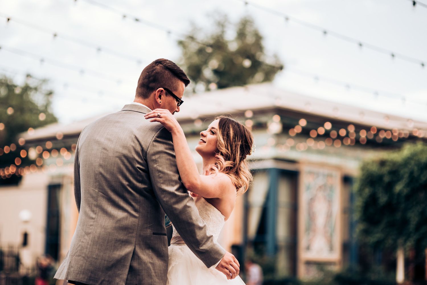 A romantic sunset wedding portrait with string lights and a rustic building in the background.