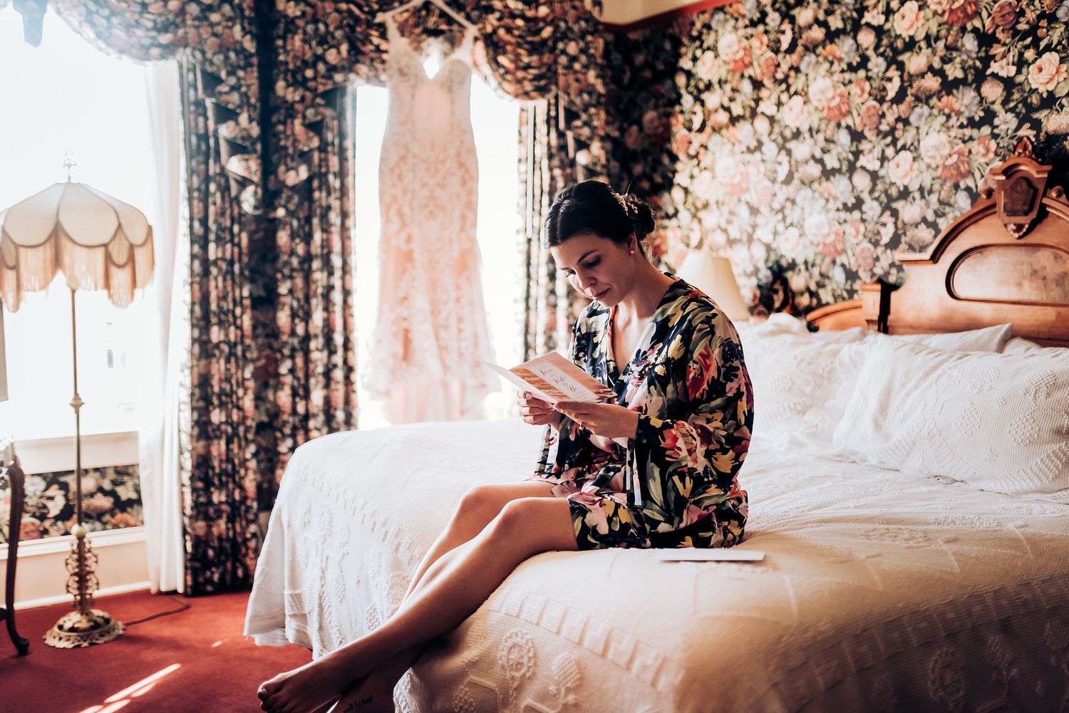 Person in floral robe sitting on bed with wedding dress hanging in background.