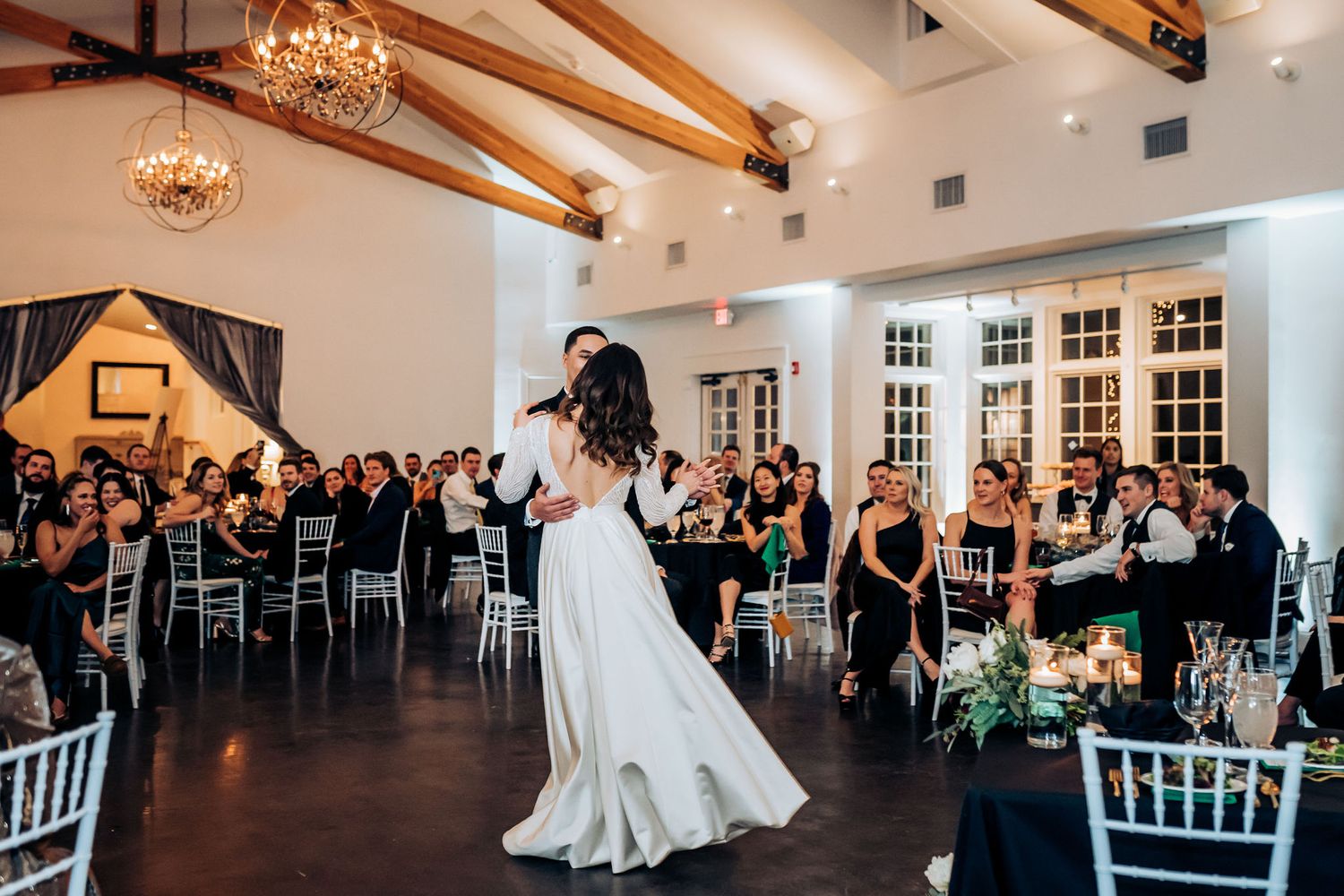Wedding couple dances in an elegant ballroom while guests watch from decorated dining tables.