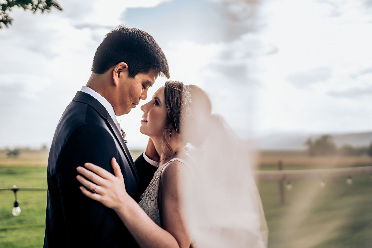 Romantic couple embrace during sunset with veil flowing in outdoor setting.