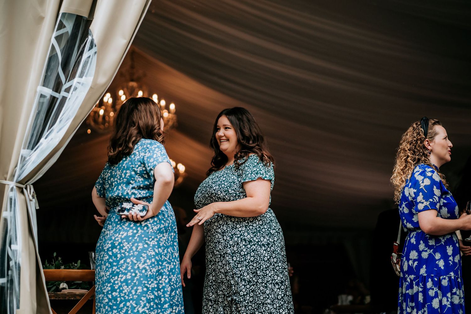 Wedding guests in floral dresses socialize inside a white tent reception.