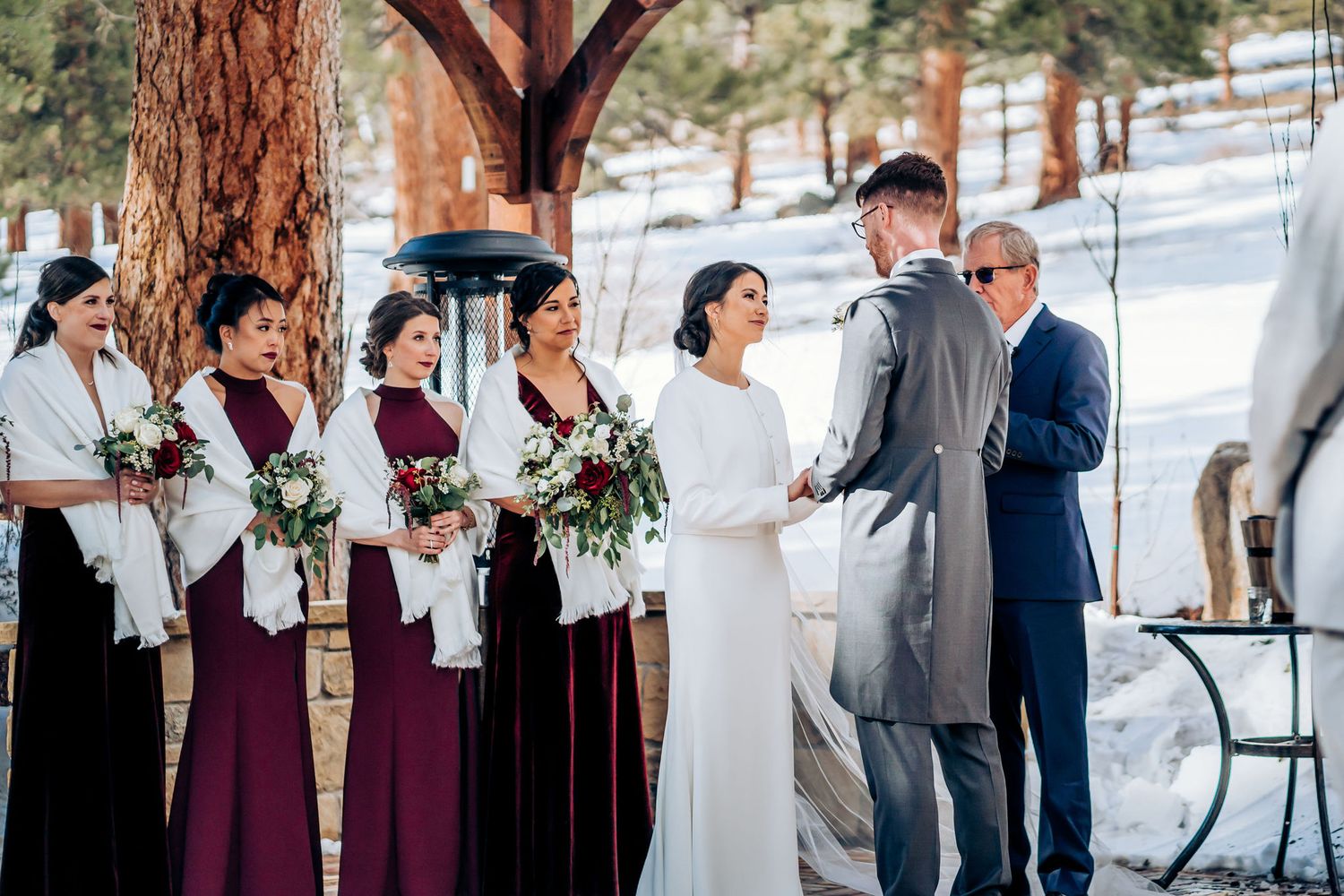 Winter wedding ceremony under wooden pergola with snow-covered landscape and burgundy bridesmaids.