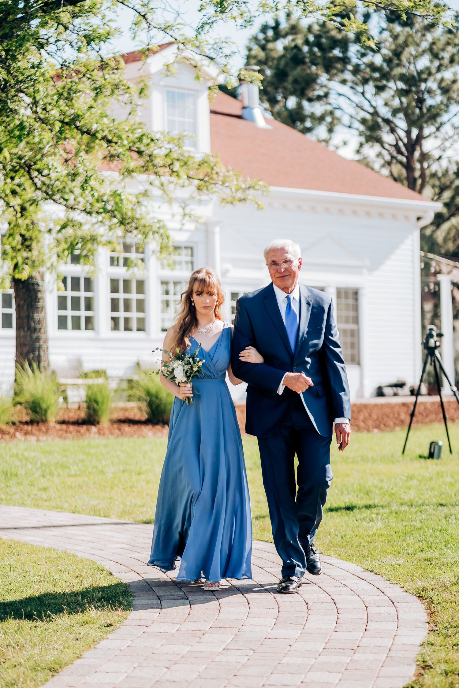 Couple walking down brick path in formal blue attire outside white house.