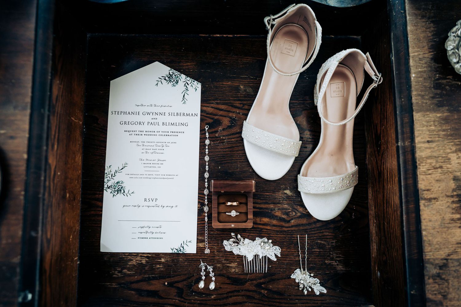 Wedding details flatlay with white shoes and invitation on dark wooden surface.
