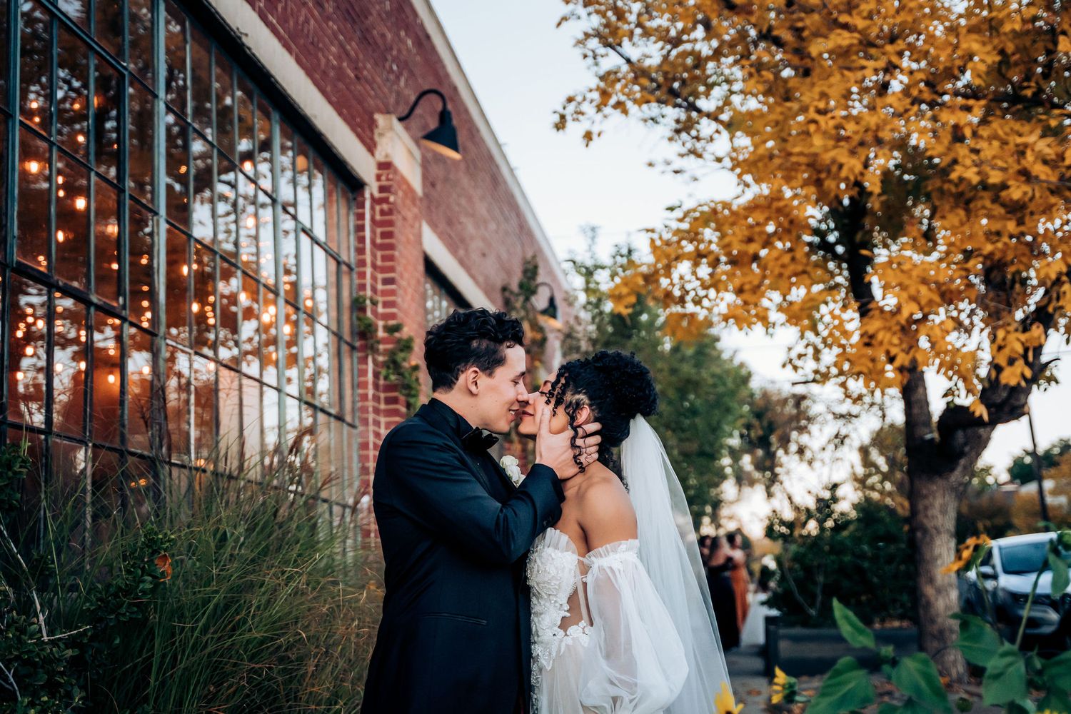 Newlyweds share a tender moment outside a brick building with autumn foliage and string lights.