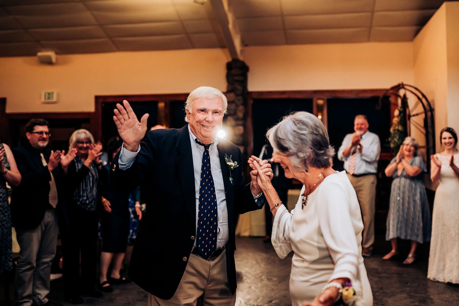 An elderly couple dancing joyfully at a wedding reception while other guests applaud.