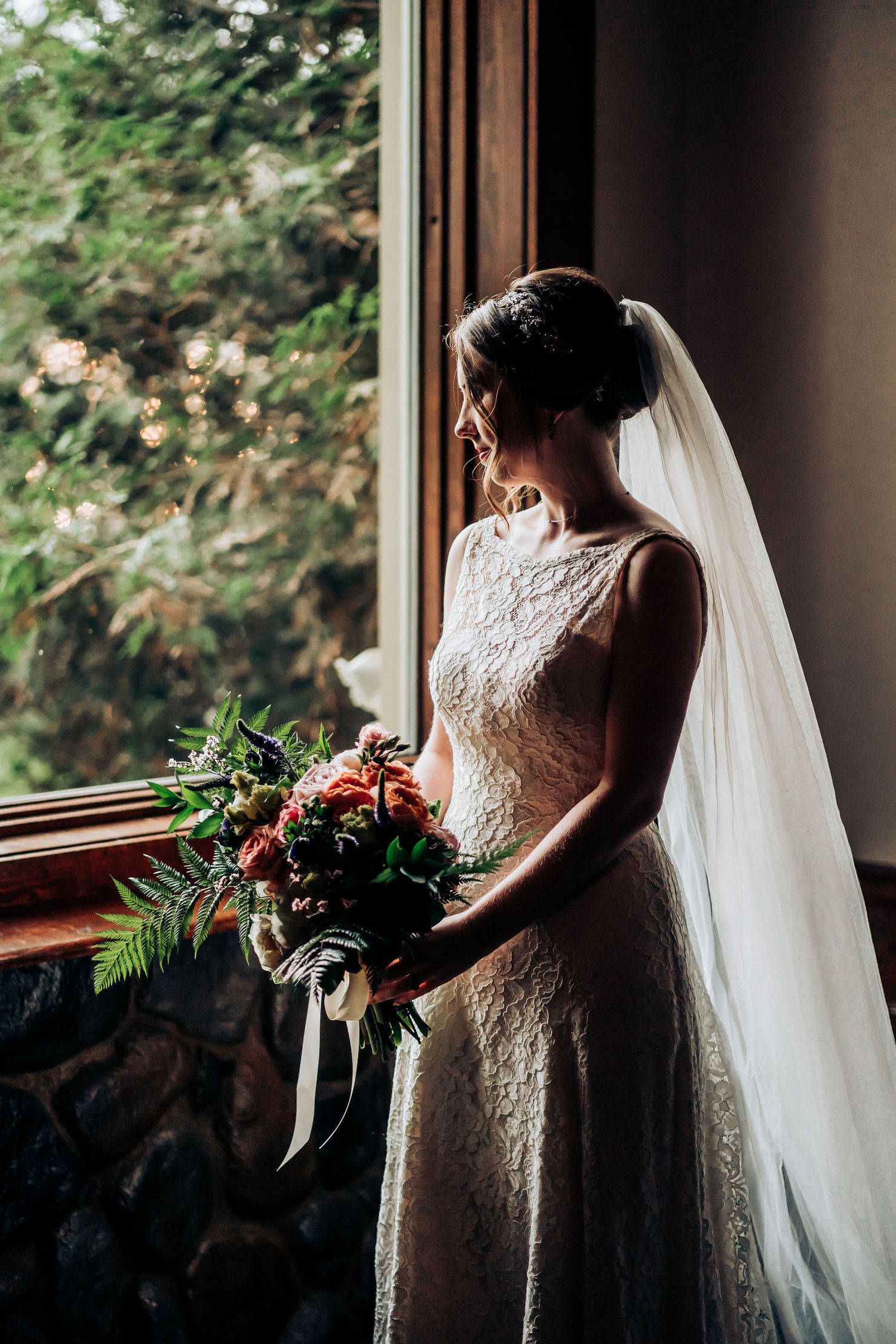 A bride in lace dress holds a bouquet while gazing out a window in soft natural light.