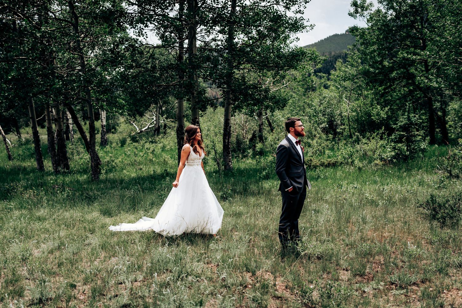 A couple in wedding attire stand apart in a scenic mountain meadow surrounded by pine trees.