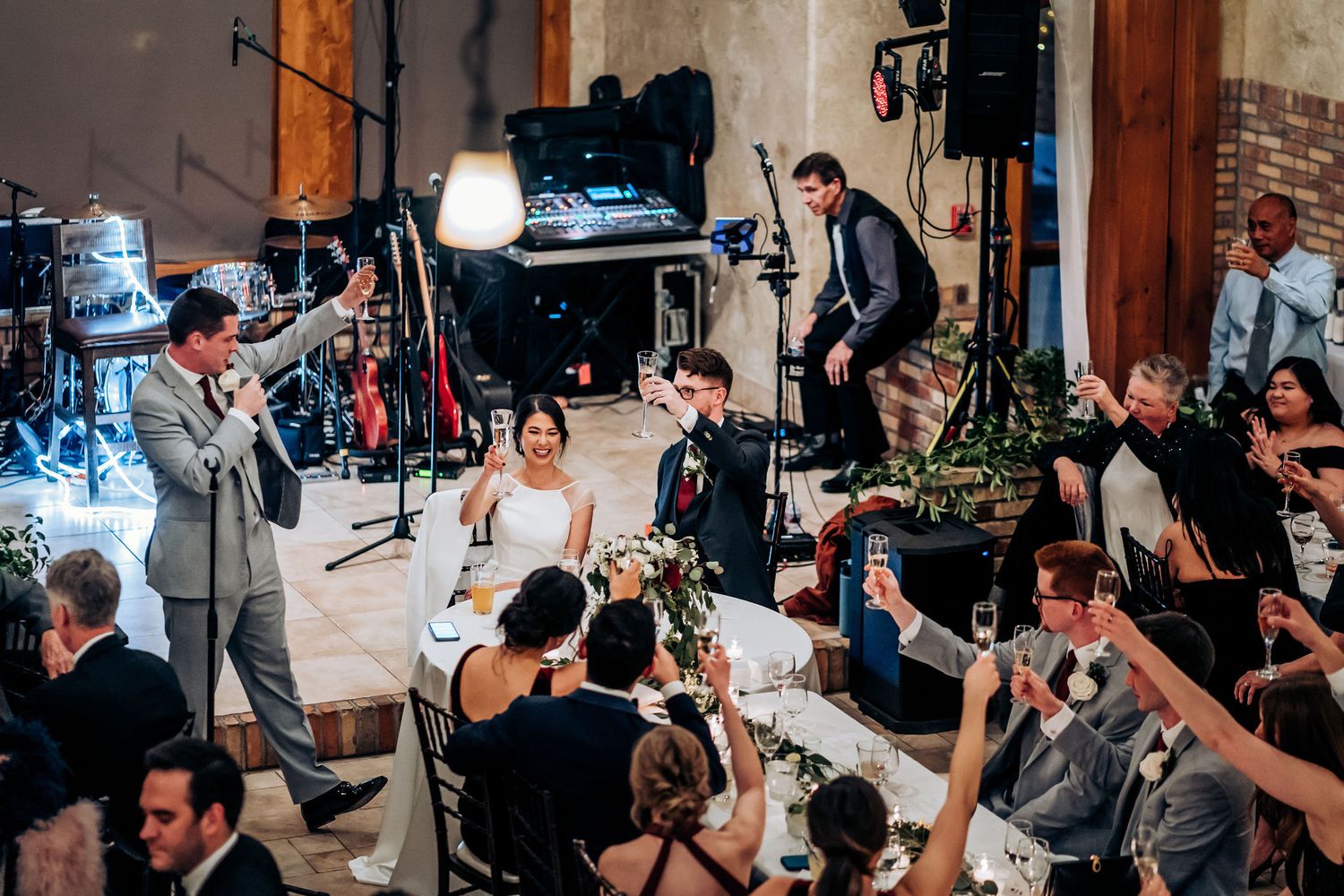 Wedding reception guests watching and listening to speeches in a rustic venue with professional lighting setup.
