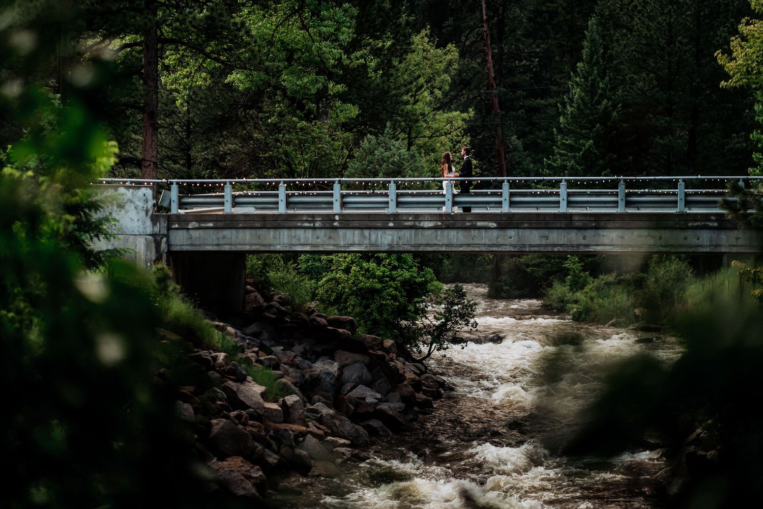 A distant view of a couple standing on a wooden bridge over a rushing stream in a forest.