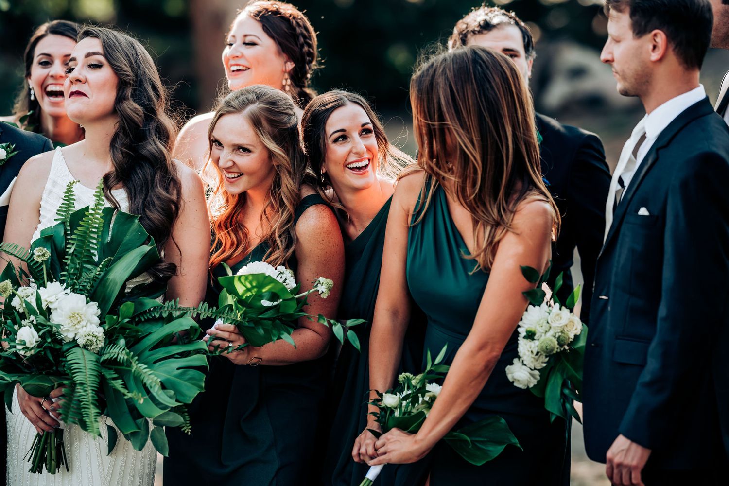 Bridesmaids in emerald green dresses smile while holding lush green and white bouquets.