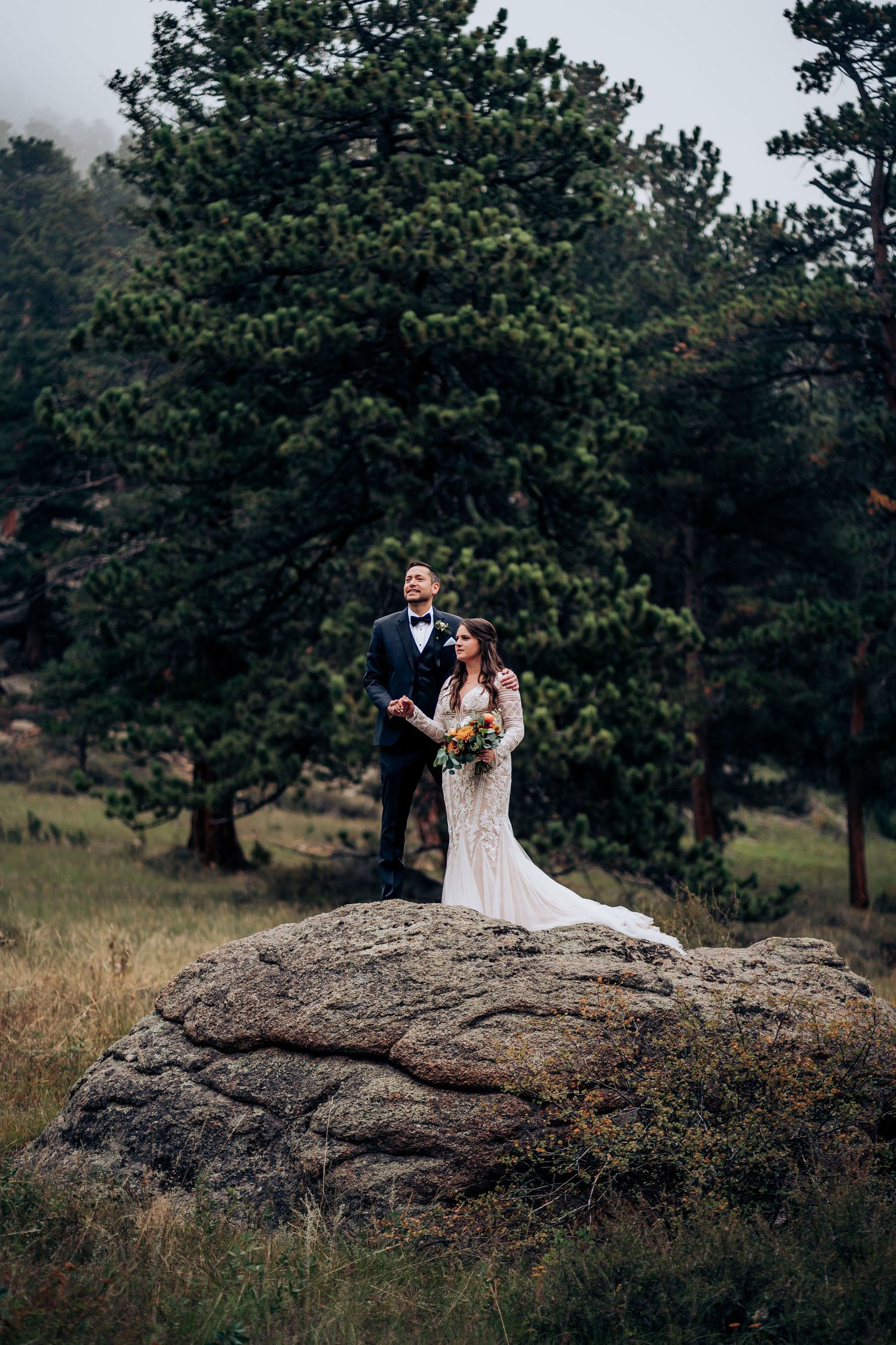 A couple stands atop a large boulder in wedding attire surrounded by pine trees.