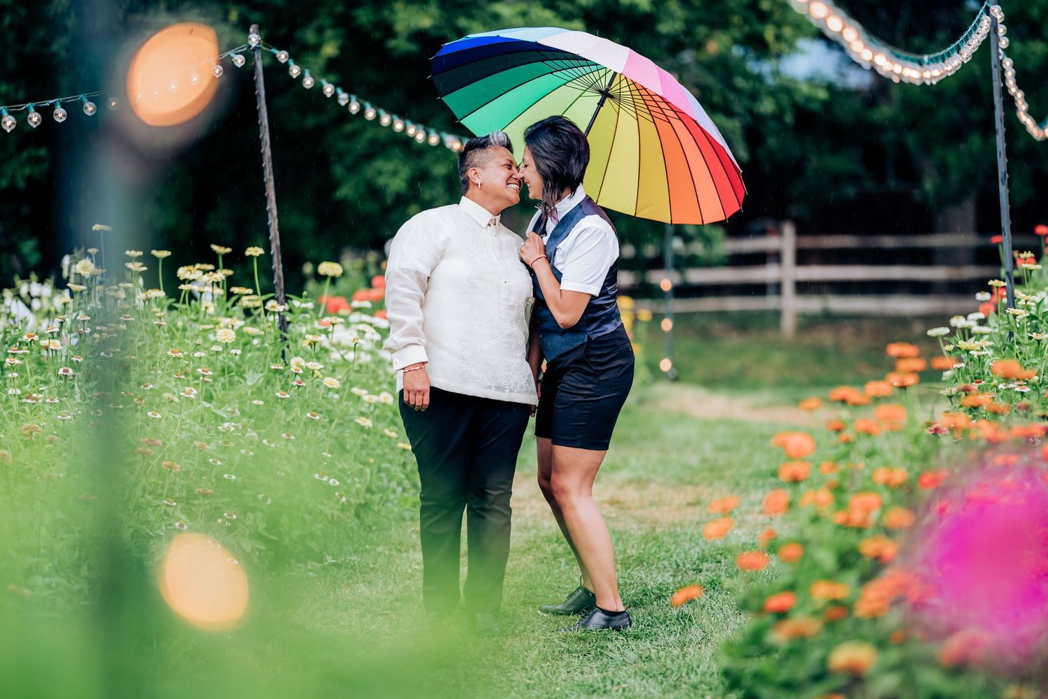 A couple shares an intimate moment under a rainbow umbrella in a flower garden.