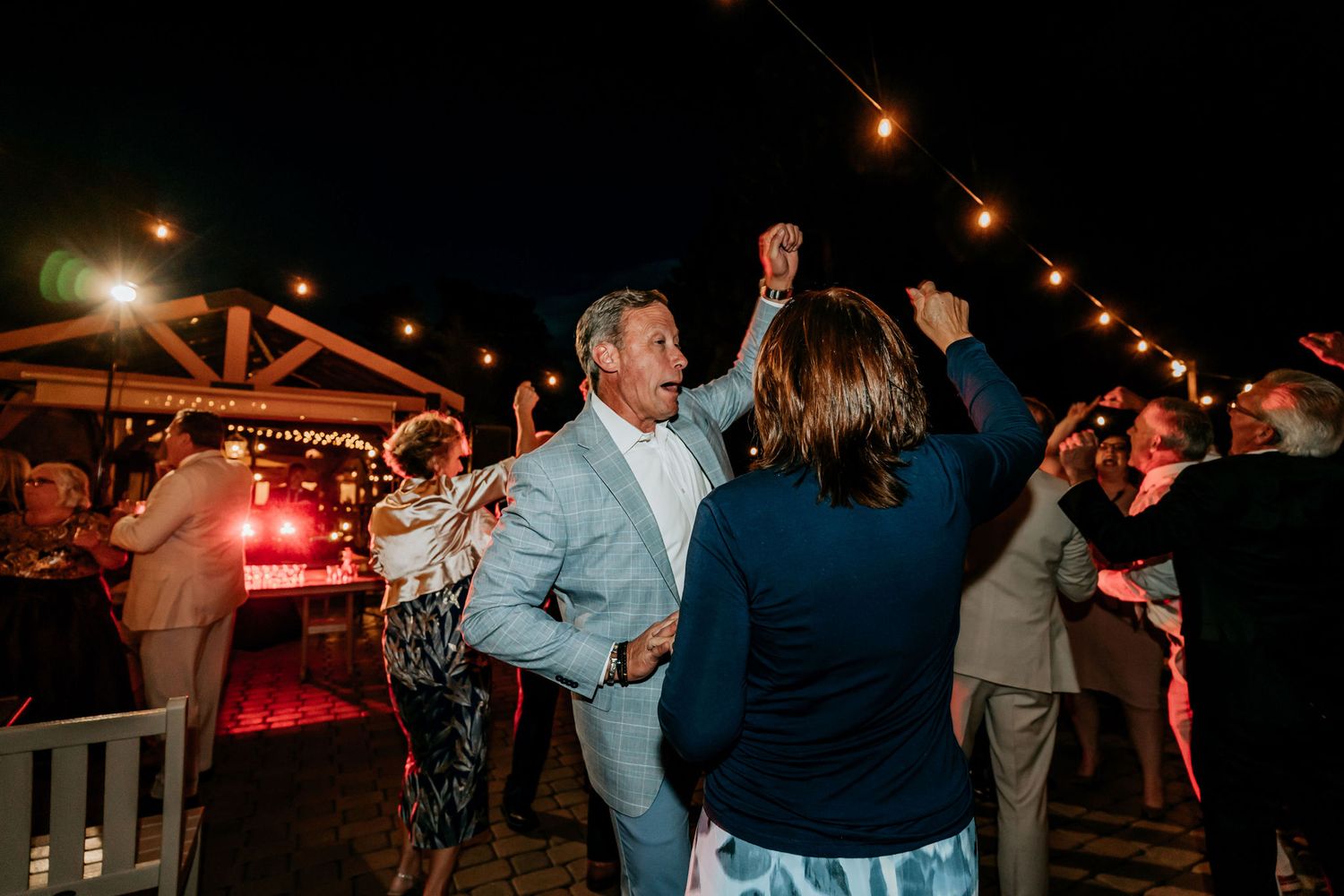 Wedding guests dance joyfully under twinkling string lights at an outdoor evening reception.