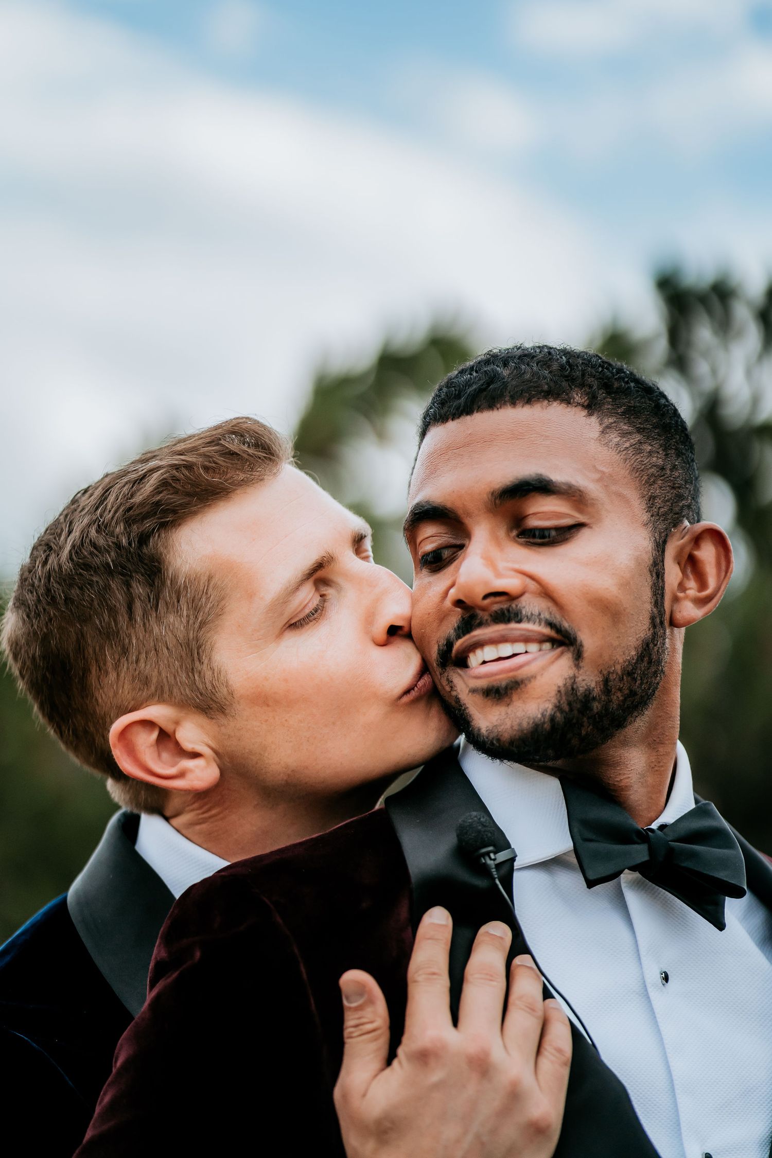 A tender moment between two grooms in formal attire sharing an intimate kiss against a blurred natural backdrop.