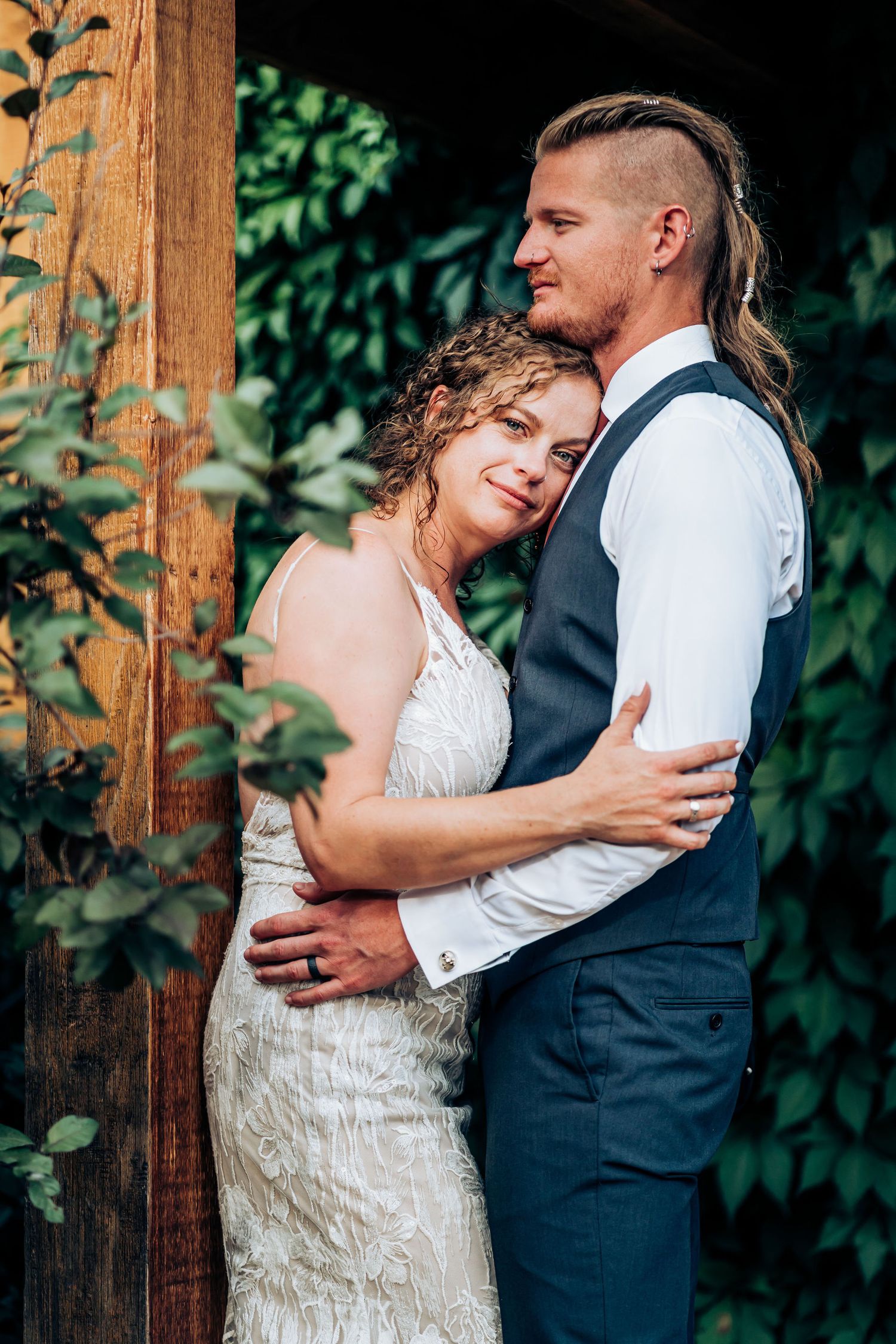 Newlyweds share intimate moment against wooden pillar with greenery.