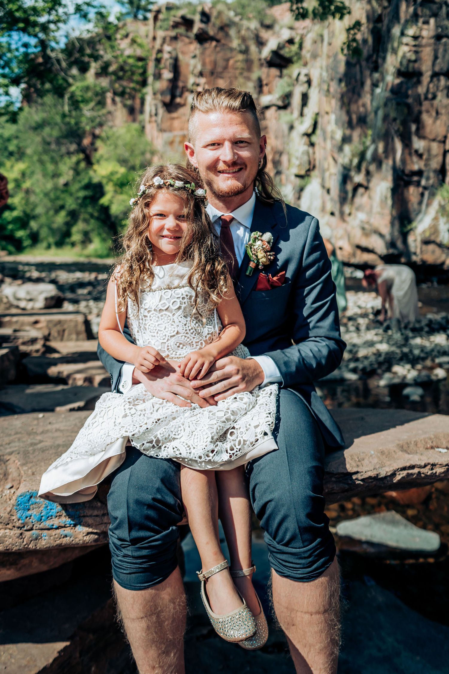 A wedding photo of a pair in a white dress and navy suit sitting on rocks near a cliff face.