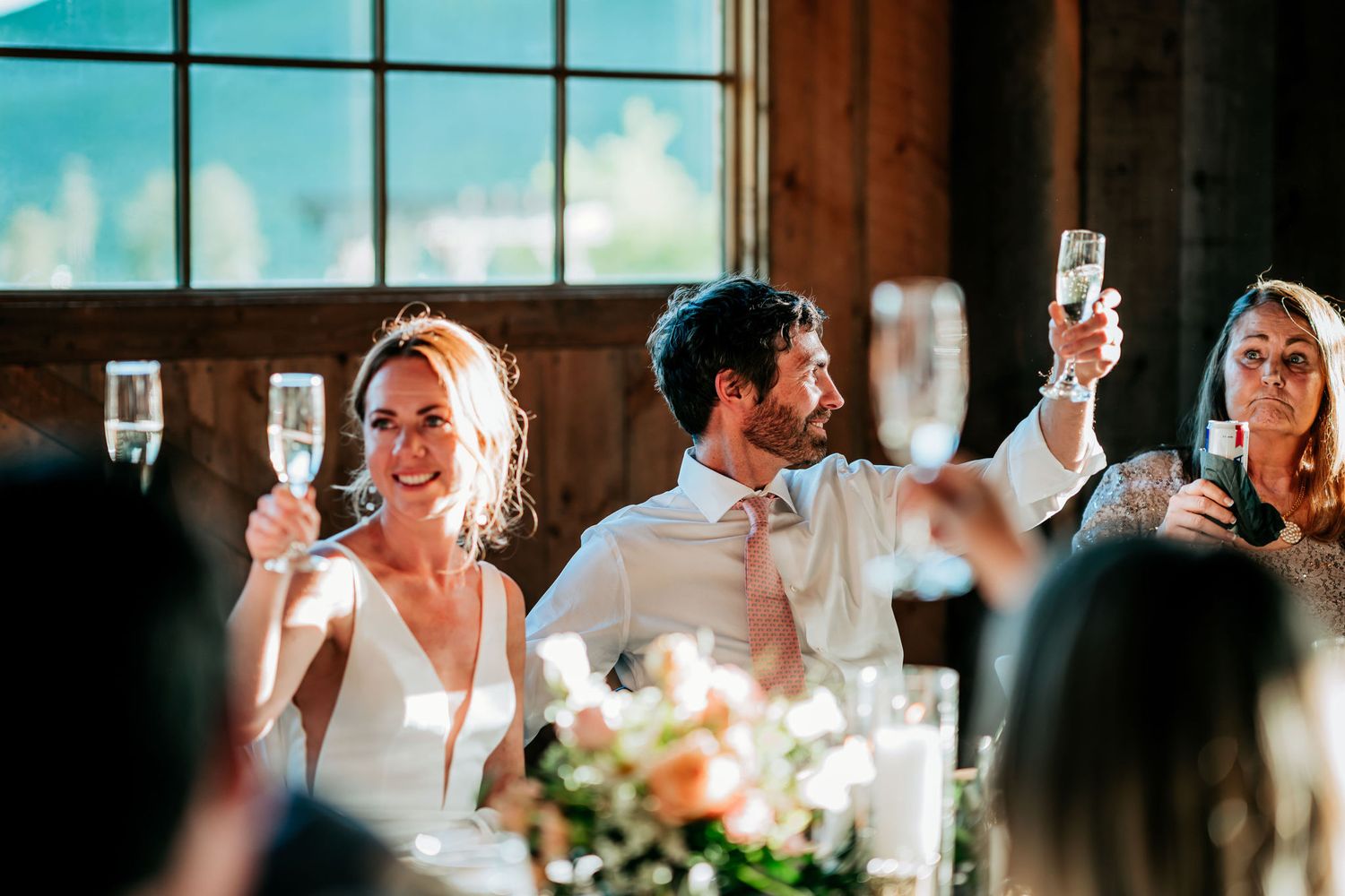 Guests raise champagne glasses in celebration during a wedding reception in a sunlit rustic venue.