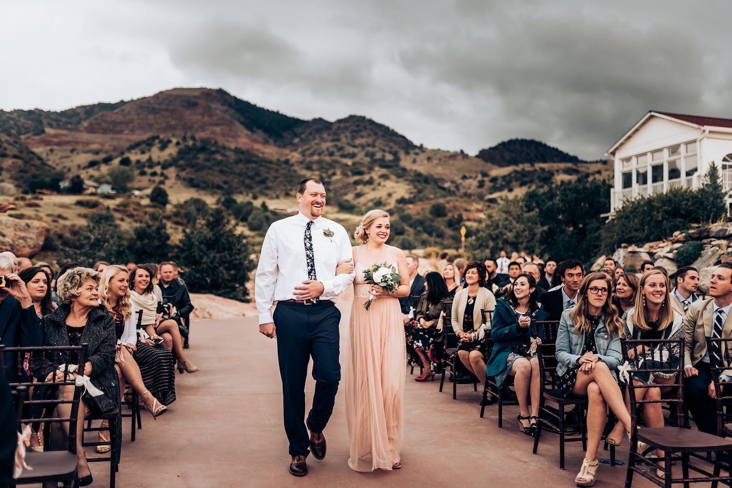 Wedding processional on an outdoor terrace with mountain views and seated guests.