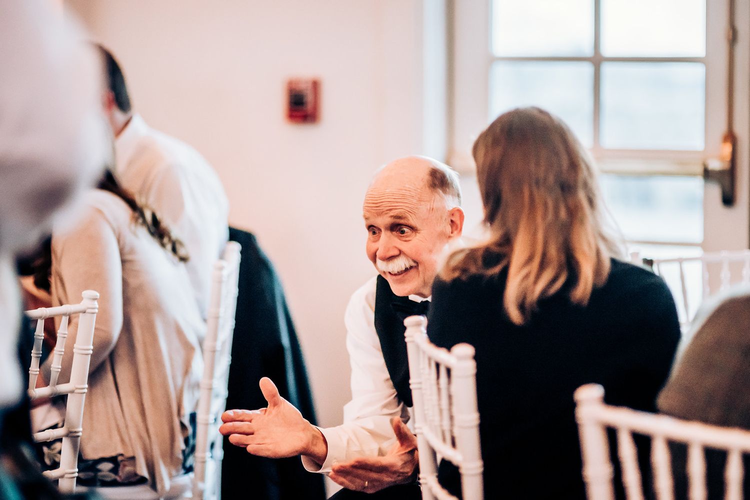 Candid indoor reception photo showing guests seated at white chairs engaged in conversation.