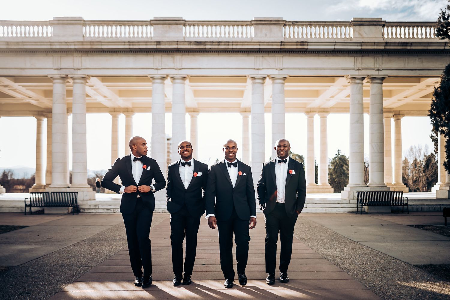 Four groomsmen walking together under elegant white colonnade in formal attire.