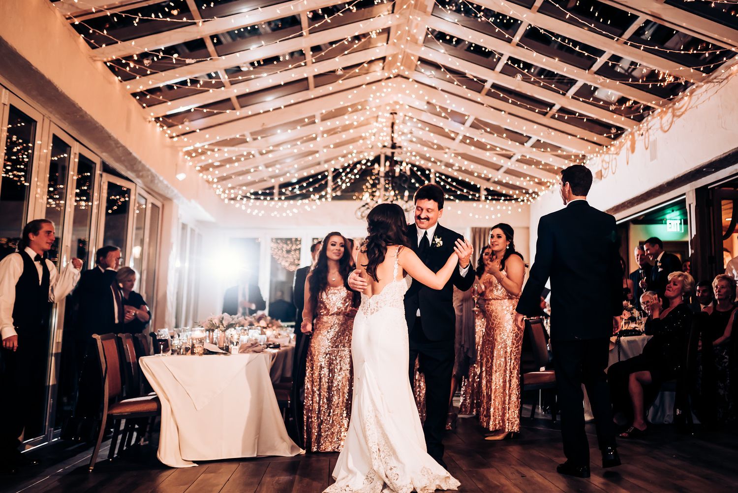 Couple dances as bridesmaids in rose gold dresses watch during wedding reception under fairy lights.