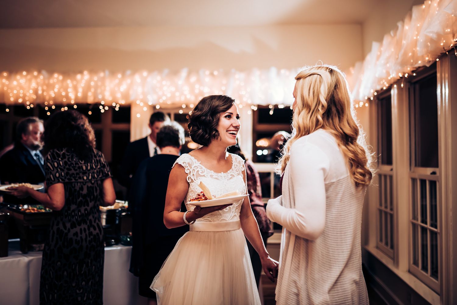 Two guests engaged in animated conversation at an evening wedding reception with twinkling lights overhead.