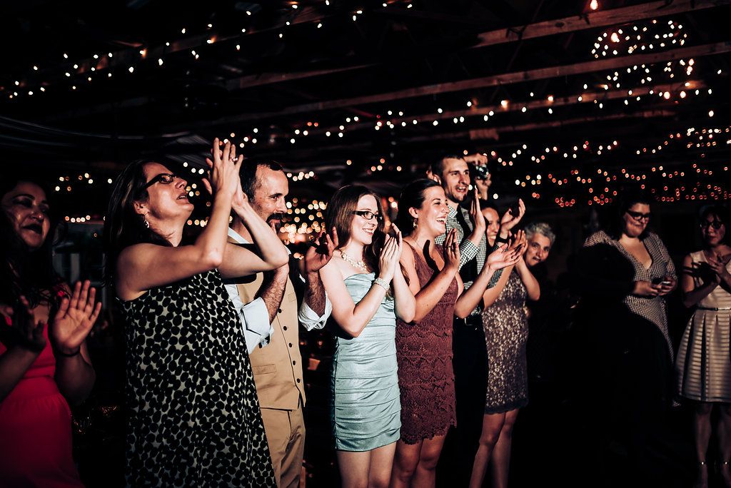 Crowd of party guests applaud during an indoor celebration with string lights overhead.