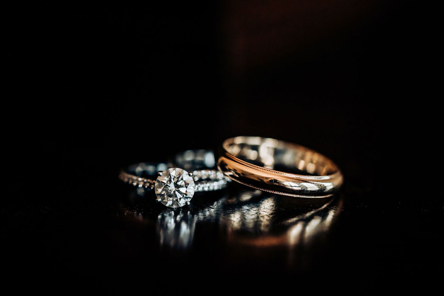 Wedding rings including diamond engagement ring and gold band reflecting on dark surface.